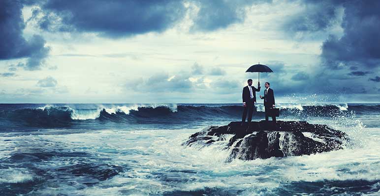 Two businessmen standing under an umbrella on a lone rock in the ocean with waves crashing around them.
