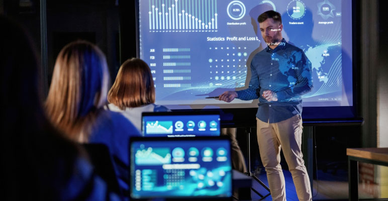 Photograph of man standing in front of three people sitting at desks with laptops while he is pointing at a bar graph on a large screen.